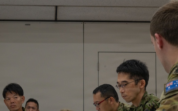 A military meeting or briefing session involving personnel from at least two nations — Australia and Japan — seated around a table in an indoor facility. The Australian soldier is identifiable by the distinctive Southern Cross flag patch on his multicam uniform, while the other personnel wear Japanese Ground Self-Defense Force camouflage uniforms. Slouch hats with badge insignia are placed on the table, indicating an Australian Army presence.