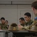A military meeting or briefing session involving personnel from at least two nations — Australia and Japan — seated around a table in an indoor facility. The Australian soldier is identifiable by the distinctive Southern Cross flag patch on his multicam uniform, while the other personnel wear Japanese Ground Self-Defense Force camouflage uniforms. Slouch hats with badge insignia are placed on the table, indicating an Australian Army presence.