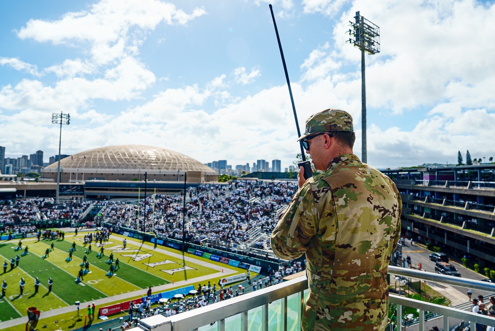 A military personnel in OCP (Operational Camouflage Pattern) uniform is standing on an elevated vantage point overlooking a packed American football stadium, speaking into a handheld radio. The stadium field displays a large 'H' logo, suggesting this is the University of Hawaii's Clarence T.C. Ching Athletics Complex (Aloha Stadium area). A distinctive domed structure and urban skyline are visible in the background, consistent with Honolulu, Hawaii.