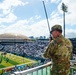A military personnel in OCP (Operational Camouflage Pattern) uniform is standing on an elevated vantage point overlooking a packed American football stadium, speaking into a handheld radio. The stadium field displays a large 'H' logo, suggesting this is the University of Hawaii's Clarence T.C. Ching Athletics Complex (Aloha Stadium area). A distinctive domed structure and urban skyline are visible in the background, consistent with Honolulu, Hawaii.