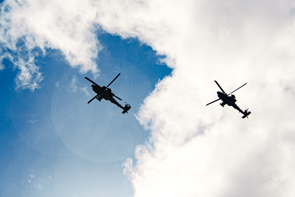 Two military attack helicopters are photographed in silhouette against a partly cloudy blue sky, shot from a low ground angle looking upward. The helicopters appear to be flying in formation or close proximity to one another. The backlighting creates strong dark silhouettes, making detailed identification challenging but the tandem-rotor attack helicopter profile is clearly distinguishable.