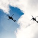 Two military attack helicopters are photographed in silhouette against a partly cloudy blue sky, shot from a low ground angle looking upward. The helicopters appear to be flying in formation or close proximity to one another. The backlighting creates strong dark silhouettes, making detailed identification challenging but the tandem-rotor attack helicopter profile is clearly distinguishable.