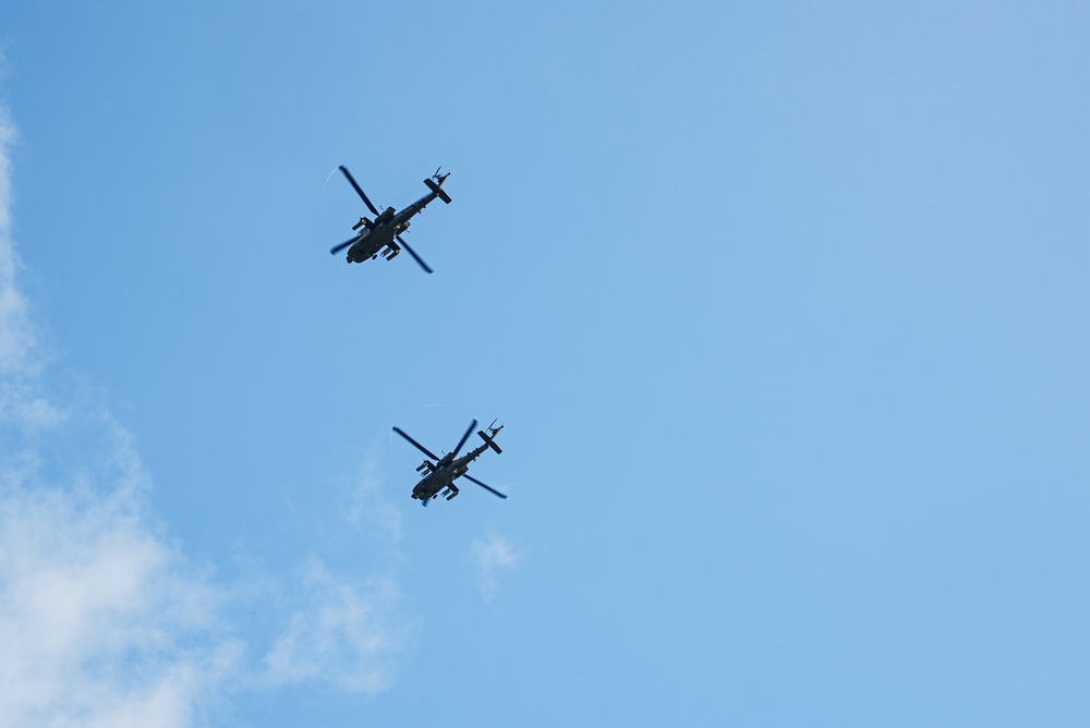 Two military attack helicopters are flying in formation against a clear blue sky with minimal cloud cover. The helicopters appear as dark silhouettes photographed from below, showing their underside profiles. Their tandem-seat, narrow-fuselage design is consistent with attack helicopter configurations.