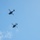 Two military attack helicopters are flying in formation against a clear blue sky with minimal cloud cover. The helicopters appear as dark silhouettes photographed from below, showing their underside profiles. Their tandem-seat, narrow-fuselage design is consistent with attack helicopter configurations.