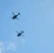 Two military attack helicopters are flying in formation against a clear blue sky with minimal cloud cover. The helicopters appear as dark silhouettes photographed from below, showing their underside profiles. Their tandem-seat, narrow-fuselage design is consistent with attack helicopter configurations.