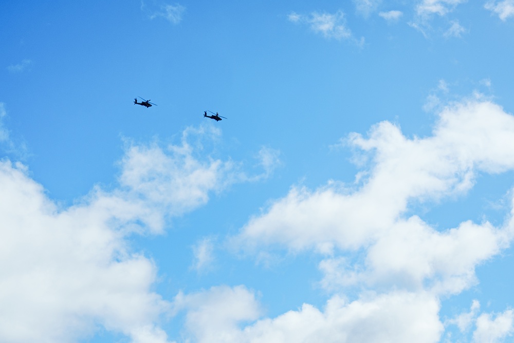 The image shows two helicopters flying in formation against a bright blue sky with scattered white cumulus clouds. The helicopters appear as dark silhouettes, suggesting they are at a considerable altitude. Their profile shapes suggest they may be military-type rotary-wing aircraft.