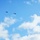 The image shows two helicopters flying in formation against a bright blue sky with scattered white cumulus clouds. The helicopters appear as dark silhouettes, suggesting they are at a considerable altitude. Their profile shapes suggest they may be military-type rotary-wing aircraft.