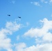 The image shows two helicopters flying in formation against a bright blue sky with scattered white cumulus clouds. The helicopters appear as dark silhouettes, suggesting they are at a considerable altitude. Their profile shapes suggest they may be military-type rotary-wing aircraft.