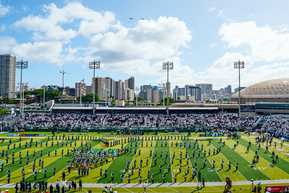 A pre-game or halftime ceremony is taking place at an outdoor American football stadium, featuring a large marching band formation on the field along with an American flag display. Military helicopters are conducting a flyover above the stadium. The venue appears to be located in an urban setting, likely Honolulu, Hawaii, based on the distinctive skyline and the domed arena visible in the background.