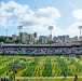 A pre-game or halftime ceremony is taking place at an outdoor American football stadium, featuring a large marching band formation on the field along with an American flag display. Military helicopters are conducting a flyover above the stadium. The venue appears to be located in an urban setting, likely Honolulu, Hawaii, based on the distinctive skyline and the domed arena visible in the background.