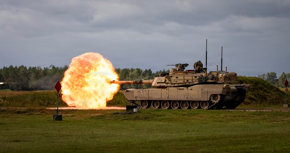 An M1 Abrams main battle tank is captured mid-fire on a training range, with a large muzzle blast fireball visible at the end of the gun barrel. The tank appears to be in a desert tan paint scheme, positioned on a grassy field with a tree line and overcast sky in the background. Red diamond-shaped range markers are visible to the left of the tank.