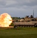An M1 Abrams main battle tank is captured mid-fire on a training range, with a large muzzle blast fireball visible at the end of the gun barrel. The tank appears to be in a desert tan paint scheme, positioned on a grassy field with a tree line and overcast sky in the background. Red diamond-shaped range markers are visible to the left of the tank.