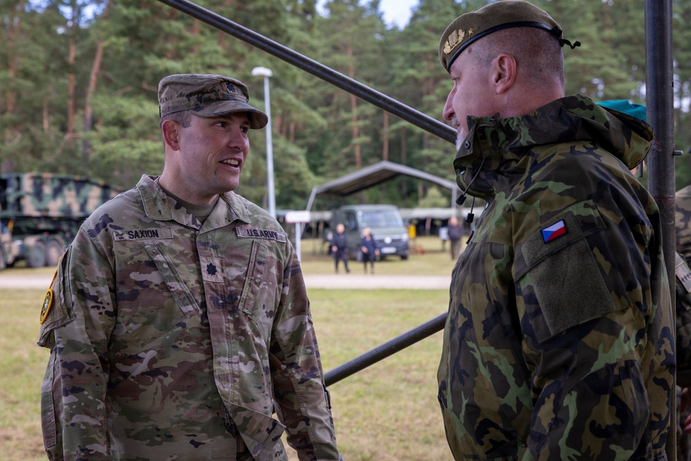 Two military personnel are engaged in conversation at what appears to be an outdoor military installation or exercise area. The soldier on the left wears U.S. Army OCP (Operational Camouflage Pattern) uniform with the name tape 'SAXION' and lieutenant colonel oak leaf insignia, while the soldier on the right wears a Czech military uniform identifiable by the Czech Republic flag patch. The setting includes military vehicles, equipment, and a forested background consistent with a joint NATO exercise or bilateral military engagement.