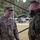 Two military personnel are engaged in conversation at what appears to be an outdoor military installation or exercise area. The soldier on the left wears U.S. Army OCP (Operational Camouflage Pattern) uniform with the name tape 'SAXION' and lieutenant colonel oak leaf insignia, while the soldier on the right wears a Czech military uniform identifiable by the Czech Republic flag patch. The setting includes military vehicles, equipment, and a forested background consistent with a joint NATO exercise or bilateral military engagement.