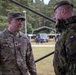 Two military personnel are engaged in conversation at what appears to be an outdoor military installation or exercise area. The soldier on the left wears U.S. Army OCP (Operational Camouflage Pattern) uniform with the name tape 'SAXION' and lieutenant colonel oak leaf insignia, while the soldier on the right wears a Czech military uniform identifiable by the Czech Republic flag patch. The setting includes military vehicles, equipment, and a forested background consistent with a joint NATO exercise or bilateral military engagement.