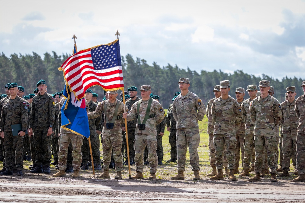 A military formation ceremony featuring U.S. Army soldiers in MultiCam/OCP uniforms standing at attention on an open field. Two color guard soldiers are holding the American flag and what appears to be a unit guidon. Personnel wearing green berets, likely from a different allied nation, are visible on the left side of the formation.
