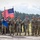 A military formation ceremony featuring U.S. Army soldiers in MultiCam/OCP uniforms standing at attention on an open field. Two color guard soldiers are holding the American flag and what appears to be a unit guidon. Personnel wearing green berets, likely from a different allied nation, are visible on the left side of the formation.