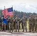 A military formation ceremony featuring U.S. Army soldiers in MultiCam/OCP uniforms standing at attention on an open field. Two color guard soldiers are holding the American flag and what appears to be a unit guidon. Personnel wearing green berets, likely from a different allied nation, are visible on the left side of the formation.