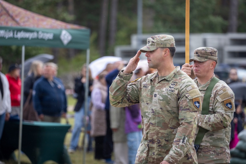 Two U.S. Army soldiers in OCP (Operational Camouflage Pattern) uniforms are participating in a military ceremony, with the foreground soldier rendering a salute. Both soldiers wear NATO patches on their left shoulders, suggesting this event is taking place at a NATO-affiliated location, likely in Poland based on the visible Polish text on the green tent. A crowd of civilians and other personnel are gathered in the background under overcast, rainy conditions.