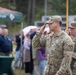 Two U.S. Army soldiers in OCP (Operational Camouflage Pattern) uniforms are participating in a military ceremony, with the foreground soldier rendering a salute. Both soldiers wear NATO patches on their left shoulders, suggesting this event is taking place at a NATO-affiliated location, likely in Poland based on the visible Polish text on the green tent. A crowd of civilians and other personnel are gathered in the background under overcast, rainy conditions.