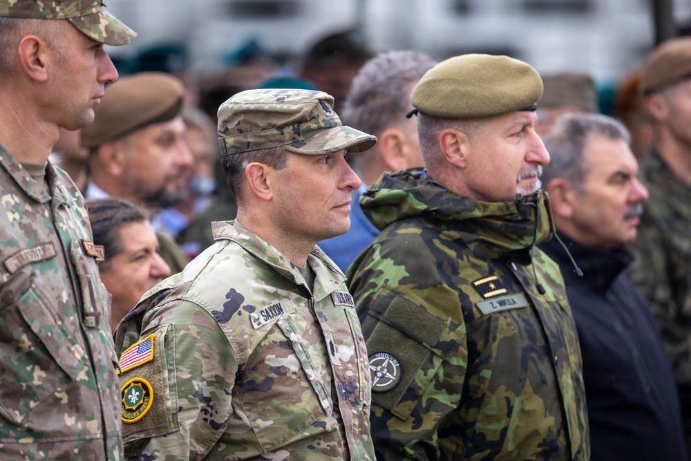 A group of military personnel from multiple nations are gathered together, likely at a multinational military event or ceremony. The foreground features a US Army soldier with visible name tape 'SAXON' and American flag patch, alongside a soldier wearing a NATO patch and name tape 'Z. MIKULA' in a different camouflage pattern. The gathering appears to be a formal or semi-formal multinational military assembly.