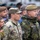 A group of military personnel from multiple nations are gathered together, likely at a multinational military event or ceremony. The foreground features a US Army soldier with visible name tape 'SAXON' and American flag patch, alongside a soldier wearing a NATO patch and name tape 'Z. MIKULA' in a different camouflage pattern. The gathering appears to be a formal or semi-formal multinational military assembly.