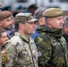 A group of military personnel from multiple nations are gathered together, likely at a multinational military event or ceremony. The foreground features a US Army soldier with visible name tape 'SAXON' and American flag patch, alongside a soldier wearing a NATO patch and name tape 'Z. MIKULA' in a different camouflage pattern. The gathering appears to be a formal or semi-formal multinational military assembly.