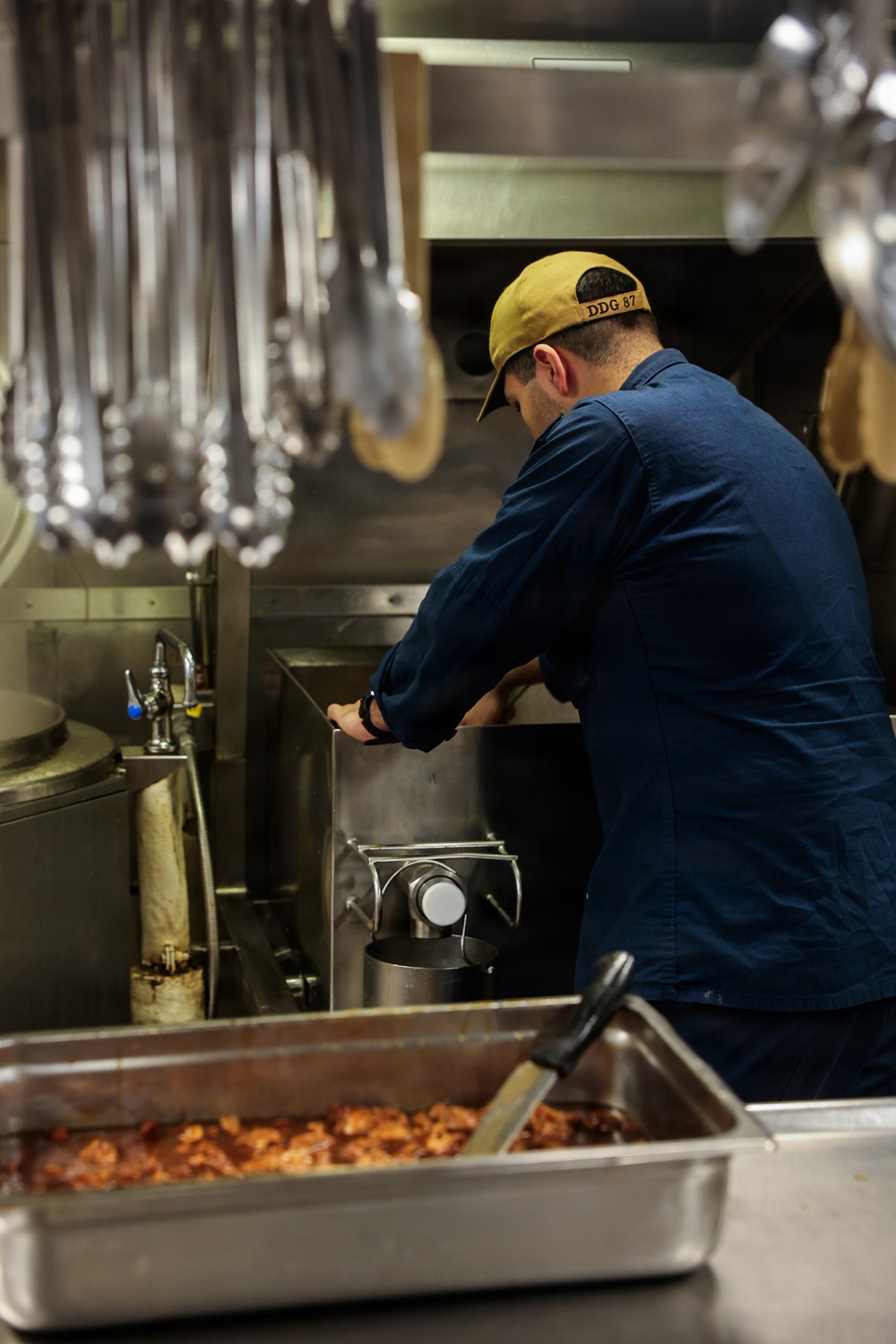 A person wearing a navy blue uniform and a yellow cap marked 'DDG 87' is working in a military ship's galley kitchen. They are leaning over large cooking equipment, likely a steam kettle or tilting braiser, in a stainless steel commercial kitchen environment. A hotel pan in the foreground contains what appears to be a cooked meat dish with sauce.