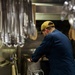 A person wearing a navy blue uniform and a yellow cap marked 'DDG 87' is working in a military ship's galley kitchen. They are leaning over large cooking equipment, likely a steam kettle or tilting braiser, in a stainless steel commercial kitchen environment. A hotel pan in the foreground contains what appears to be a cooked meat dish with sauce.