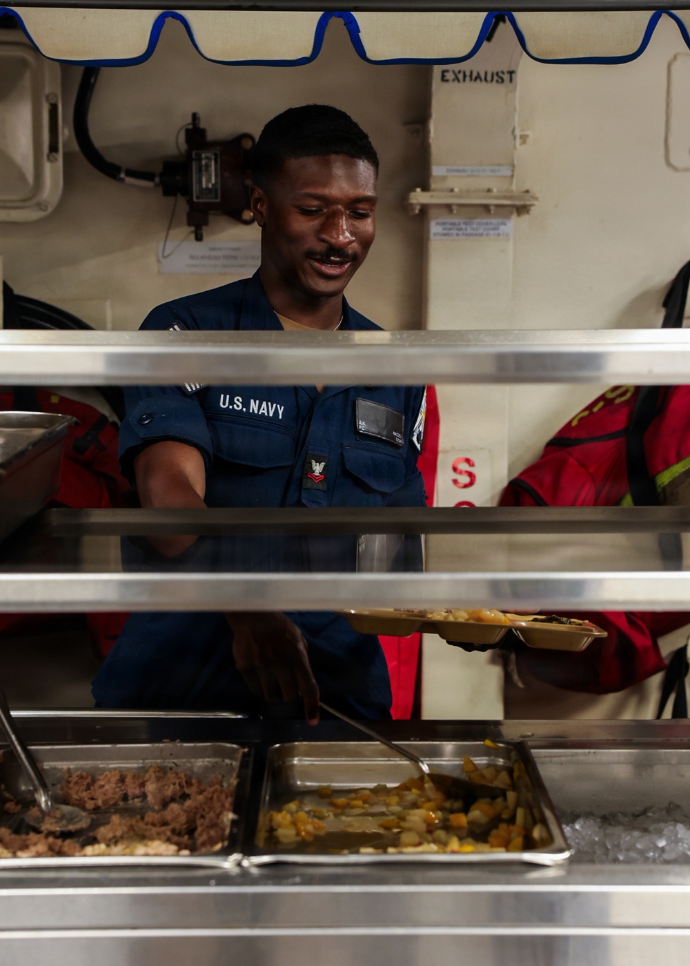 A U.S. Navy sailor in blue utility uniform is serving food at a military mess hall or galley, likely aboard a naval vessel. The sailor is smiling and holding a food tray while standing behind a stainless steel serving line with multiple food pans. The setting appears to be a shipboard galley based on the industrial equipment and 'EXHAUST' labeled ductwork visible in the background.