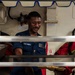 A U.S. Navy sailor in blue utility uniform is serving food at a military mess hall or galley, likely aboard a naval vessel. The sailor is smiling and holding a food tray while standing behind a stainless steel serving line with multiple food pans. The setting appears to be a shipboard galley based on the industrial equipment and 'EXHAUST' labeled ductwork visible in the background.