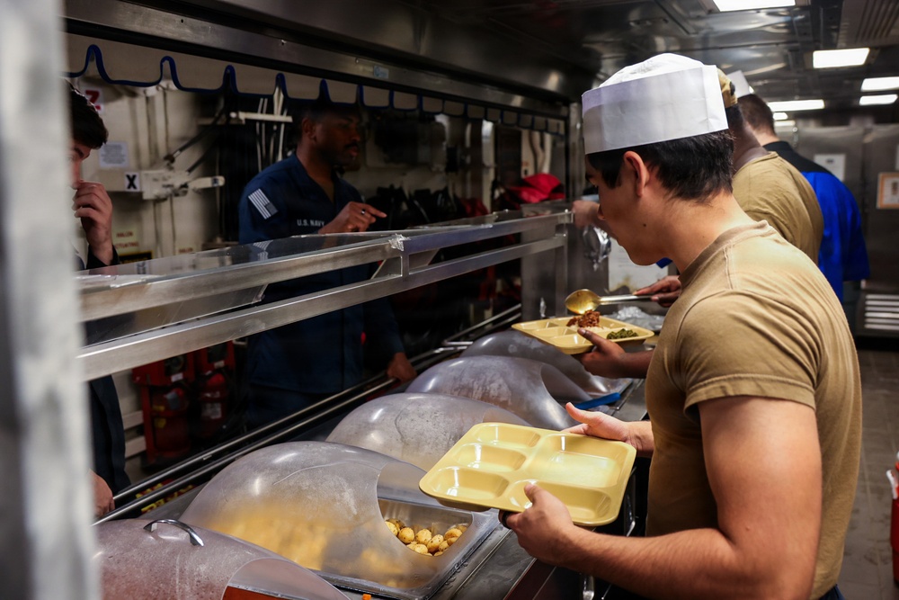 The image depicts a U.S. Navy mess hall (galley) aboard what appears to be a naval vessel, where sailors are going through a cafeteria-style food service line. A cook wearing a white chef's hat and a sailor in a blue U.S. Navy uniform are visible, with personnel holding yellow compartmentalized meal trays. The setting features stainless steel serving equipment, covered food containers, and industrial kitchen fixtures typical of military shipboard dining facilities.