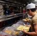 The image depicts a U.S. Navy mess hall (galley) aboard what appears to be a naval vessel, where sailors are going through a cafeteria-style food service line. A cook wearing a white chef's hat and a sailor in a blue U.S. Navy uniform are visible, with personnel holding yellow compartmentalized meal trays. The setting features stainless steel serving equipment, covered food containers, and industrial kitchen fixtures typical of military shipboard dining facilities.