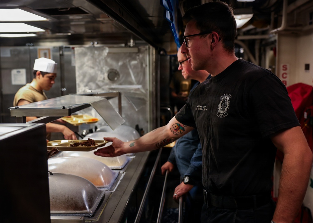 The image shows a military mess hall (galley) food service line, likely aboard a naval vessel. A sailor in a black military t-shirt with tattoos on his forearm is holding a compartmentalized food tray while being served by a cook wearing a white chef's hat. Additional personnel are visible waiting in line behind him.