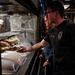 The image shows a military mess hall (galley) food service line, likely aboard a naval vessel. A sailor in a black military t-shirt with tattoos on his forearm is holding a compartmentalized food tray while being served by a cook wearing a white chef's hat. Additional personnel are visible waiting in line behind him.