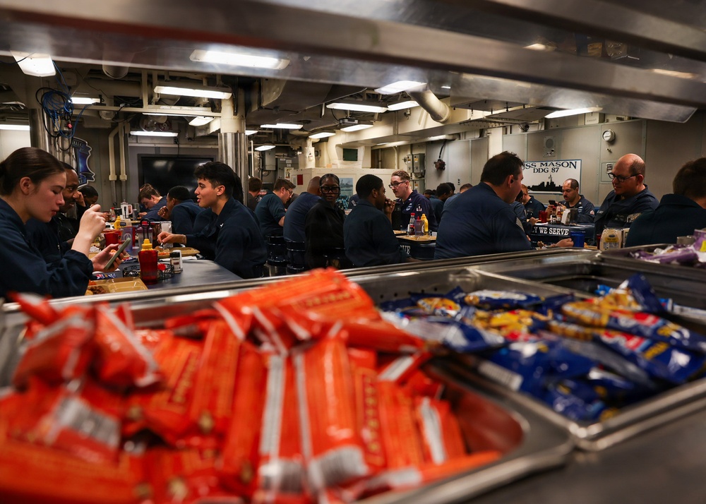 The image shows a military mess hall (galley) aboard USS Mason DDG-87, a U.S. Navy destroyer, with numerous sailors in navy blue uniforms eating a meal. The foreground features serving trays filled with packaged snacks including red and blue wrapped items. A sign reading 'USS MASON DDG 87' and another reading 'WHY WE SERVE' are visible in the background.