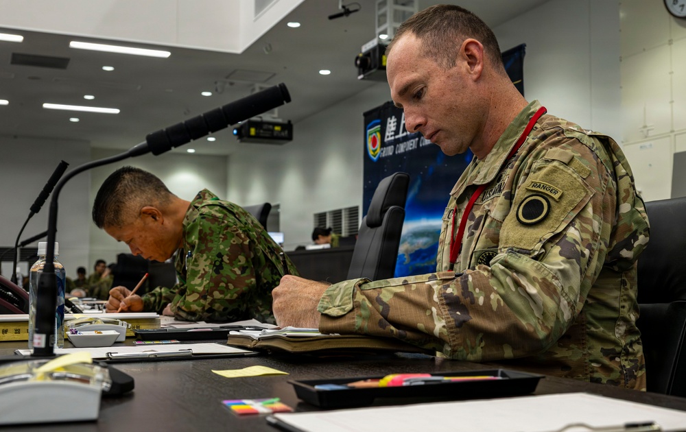 Two military personnel in different camouflage uniforms are seated at a conference table, taking notes or writing during what appears to be a joint military briefing or planning session. The soldier in the foreground wears US Army OCP (Operational Camouflage Pattern) uniform with a Ranger tab visible on the left shoulder. The setting appears to be a formal military command center or briefing room, suggesting a multinational military exercise or coordination meeting.