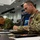 Two military personnel in different camouflage uniforms are seated at a conference table, taking notes or writing during what appears to be a joint military briefing or planning session. The soldier in the foreground wears US Army OCP (Operational Camouflage Pattern) uniform with a Ranger tab visible on the left shoulder. The setting appears to be a formal military command center or briefing room, suggesting a multinational military exercise or coordination meeting.