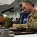 Two military personnel in different camouflage uniforms are seated at a conference table, taking notes or writing during what appears to be a joint military briefing or planning session. The soldier in the foreground wears US Army OCP (Operational Camouflage Pattern) uniform with a Ranger tab visible on the left shoulder. The setting appears to be a formal military command center or briefing room, suggesting a multinational military exercise or coordination meeting.