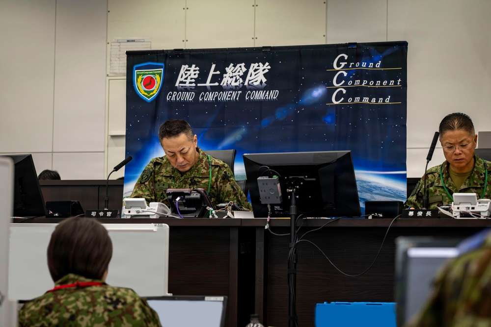 The image shows a Japan Ground Self-Defense Force (JGSDF) command center operation, with personnel seated at workstations beneath a banner reading '陸上総隊 Ground Component Command' (GCC). Two uniformed officers in camouflage uniforms are prominently visible at a command desk, with name placards indicating their roles as Commander (司令官) and Chief of Staff (幕僚長). The setting appears to be a formal military command and control exercise or operational briefing.