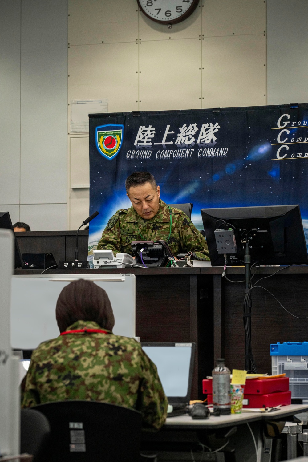 A military command center of the Japan Ground Self-Defense Force's Ground Component Command (陸上総隊) is depicted, with personnel in camouflage uniforms working at desks equipped with computers and communication equipment. A senior officer sits at a command desk in front of a large banner displaying the unit's insignia and name in both Japanese and English. The setting appears to be an operational command post during an exercise or active duty period.