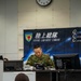 A military command center of the Japan Ground Self-Defense Force's Ground Component Command (陸上総隊) is depicted, with personnel in camouflage uniforms working at desks equipped with computers and communication equipment. A senior officer sits at a command desk in front of a large banner displaying the unit's insignia and name in both Japanese and English. The setting appears to be an operational command post during an exercise or active duty period.