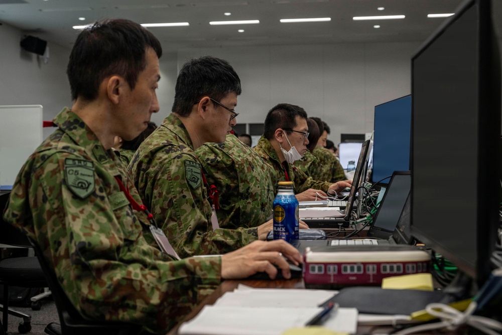Several personnel in Japanese Ground Self-Defense Force (JGSDF) camouflage uniforms are seated at workstations operating computers in what appears to be a military operations or command center. The individuals are wearing unit patches and red lanyards, suggesting a controlled-access environment. The setting resembles a joint exercise or operational planning facility.