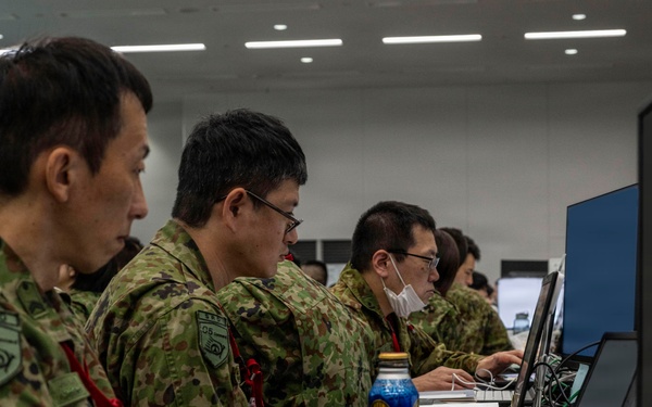 Several personnel in Japanese Ground Self-Defense Force (JGSDF) camouflage uniforms are seated at workstations operating computers in what appears to be a military operations or command center. The individuals are wearing unit patches and red lanyards, suggesting a controlled-access environment. The setting resembles a joint exercise or operational planning facility.