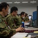 Several personnel in Japanese Ground Self-Defense Force (JGSDF) camouflage uniforms are seated at workstations operating computers in what appears to be a military operations or command center. The individuals are wearing unit patches and red lanyards, suggesting a controlled-access environment. The setting resembles a joint exercise or operational planning facility.