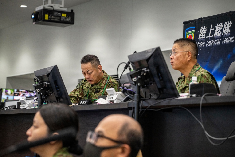 Military personnel in camouflage uniforms are working at computer workstations in what appears to be a command and control center. A banner in the background displays Japanese text '陸上総隊' (Ground Component Command) along with an English translation, indicating this is a Japan Ground Self-Defense Force facility. Multiple monitors and communication equipment are visible throughout the room.