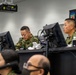 Military personnel in camouflage uniforms are working at computer workstations in what appears to be a command and control center. A banner in the background displays Japanese text '陸上総隊' (Ground Component Command) along with an English translation, indicating this is a Japan Ground Self-Defense Force facility. Multiple monitors and communication equipment are visible throughout the room.