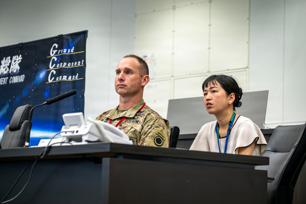 A U.S. Army soldier in multicam uniform sits alongside a civilian woman wearing a lanyard at what appears to be a press conference or briefing. Behind them is a banner reading 'Ground Component Command' with Japanese text (総隊). The setting appears to be a formal military command facility, likely in Japan.