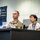 A U.S. Army soldier in multicam uniform sits alongside a civilian woman wearing a lanyard at what appears to be a press conference or briefing. Behind them is a banner reading 'Ground Component Command' with Japanese text (総隊). The setting appears to be a formal military command facility, likely in Japan.