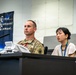 A U.S. Army soldier in multicam uniform sits alongside a civilian woman wearing a lanyard at what appears to be a press conference or briefing. Behind them is a banner reading 'Ground Component Command' with Japanese text (総隊). The setting appears to be a formal military command facility, likely in Japan.